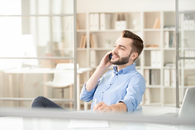 Young handsome office manager or broker with mobile phone communicating with client while sitting by workplace