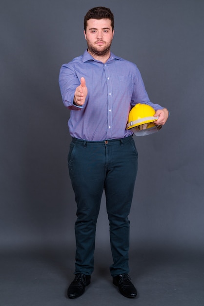 young handsome overweight bearded businessman as engineer with hardhat on gray