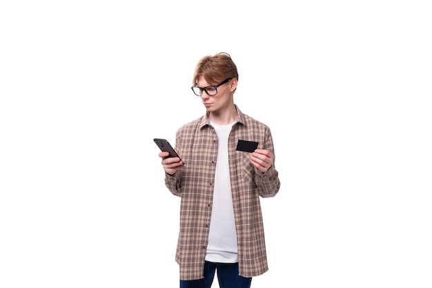 Young handsome redhead guy in a plaid shirt holds a smartphone and a credit card
