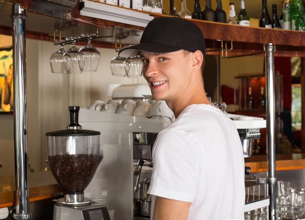 Young handsome smiling barista in white shirt and baseball hat standing bihend the counter next to the commercial grade coffeemaker. Large heavy duty espresso machine is on the background.