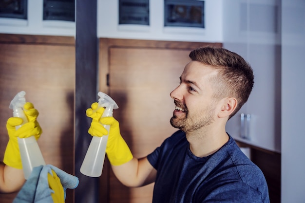 Young handsome smiling bearded man with rubber gloves spraying detergent on mirror and cleaning it with magic cloth.