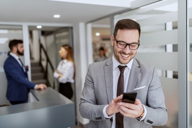 Young handsome smiling Caucasian businessman in suit talking using smart phone while standing in hall. In background his colleagues standing and chatting.