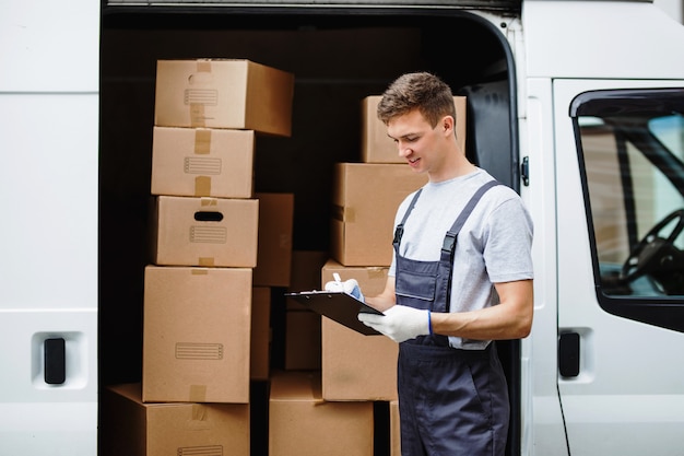 Photo a young handsome smiling worker wearing uniform is standing next to the van full of boxes holding a clipboard in his hands. house move, mover service.