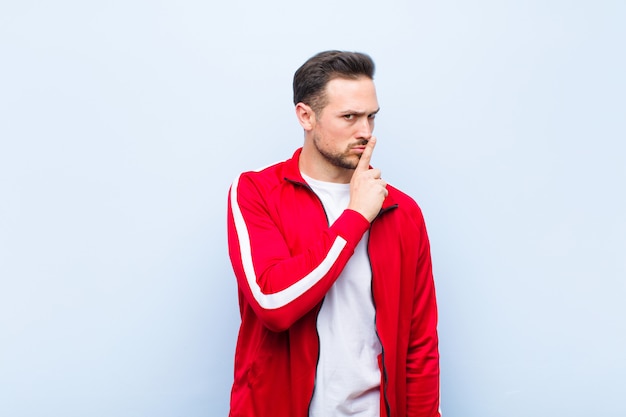 Young handsome sports man or monitor asking for silence and quiet, gesturing with finger in front of mouth, saying shh or keeping a secret against green wall