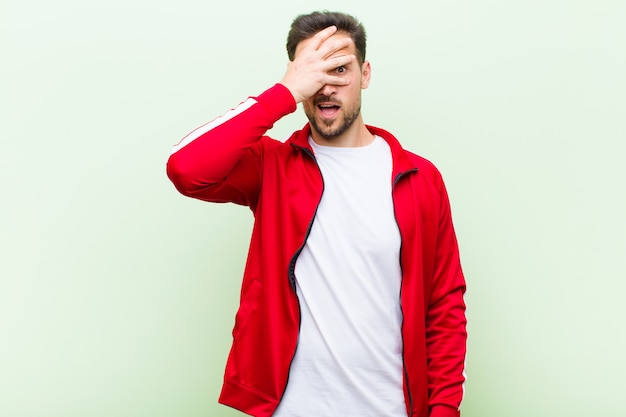 Young handsome sports man or monitor looking shocked, scared or terrified, covering face with hand and peeking between fingers against flat wall