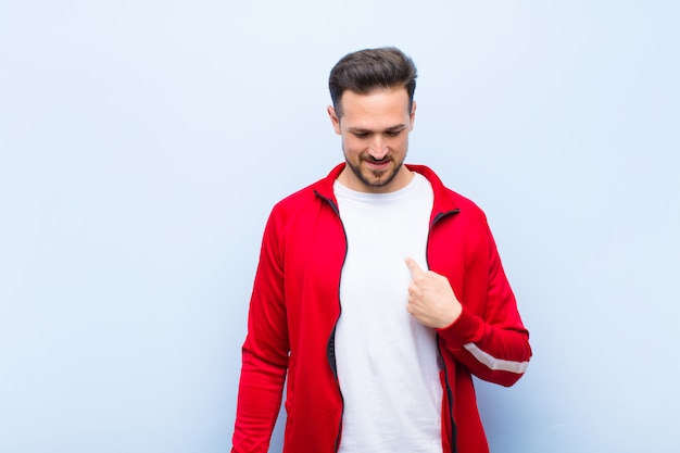 Young handsome sports man or monitor smiling cheerfully and casually, looking downwards and pointing to chest against flat wall