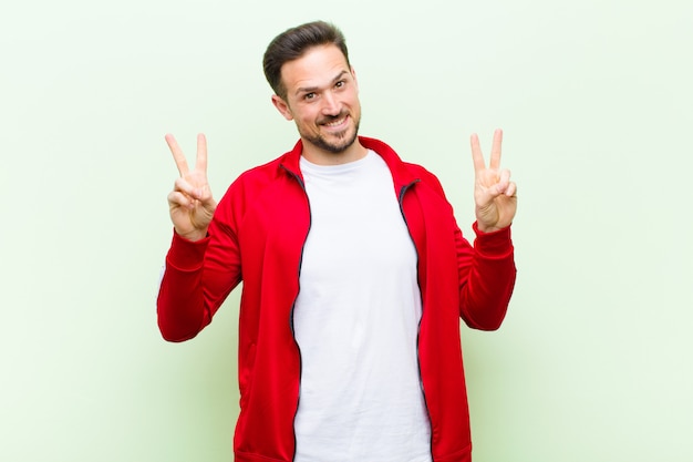 Young handsome sports man or monitor smiling and looking happy, friendly and satisfied, gesturing victory or peace with both hands against green wall