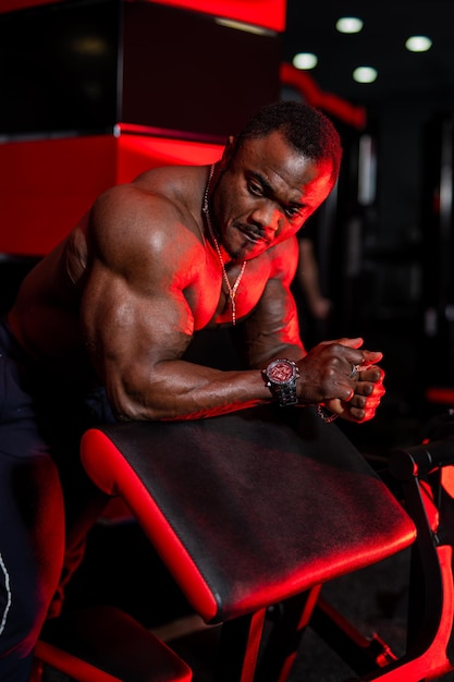 근육질의 몸을 가진 젊고 잘 생긴 스포츠맨 흑인 피트니스 남자는 스포츠 장비에 기대어 운동을 위한 체육관 배경 장비