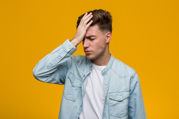 Giovane studente bello che indossa una camicia di jeans dimenticando qualcosa, schiaffeggiando la fronte con il palmo e chiudendo gli occhi.