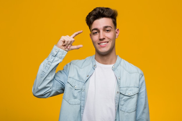 Young handsome student wearing a denim shirt holding something little with forefingers, smiling and confident.