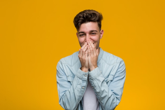 Young handsome student wearing a denim shirt laughing about something, covering mouth with hands.