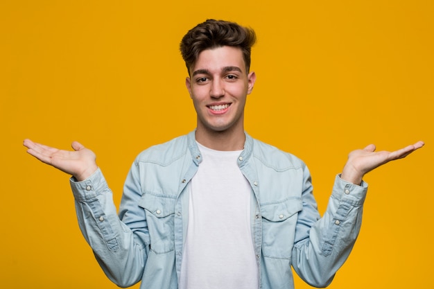 Young handsome student wearing a denim shirt makes scale with arms, feels happy and confident.