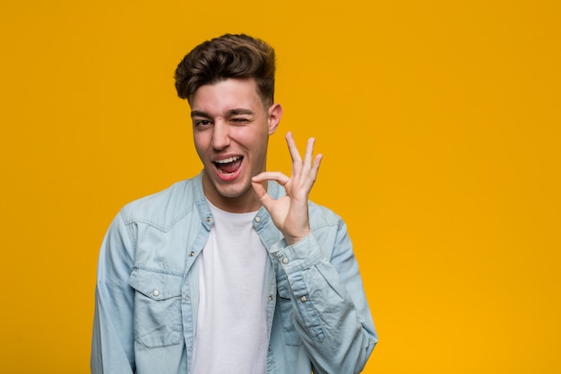 Young handsome student wearing a denim shirt winks an eye and holds an okay gesture with hand.