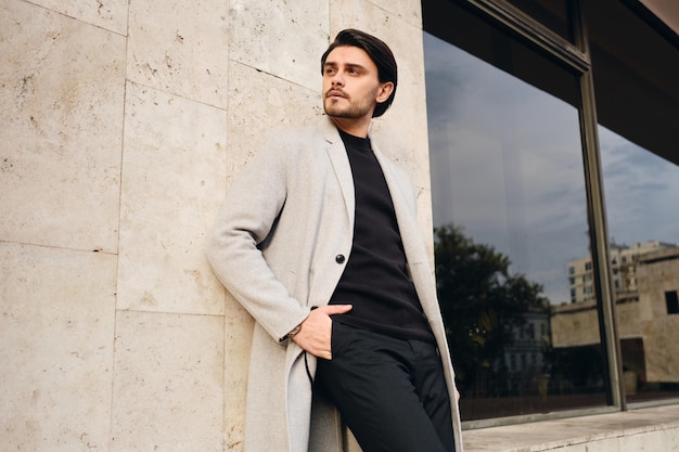 Young handsome stylish man in coat with hands in pockets intently looking away outdoor