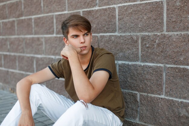 Giovane uomo alla moda bello con i capelli in una maglietta classica che si siede vicino al muro in una giornata estiva