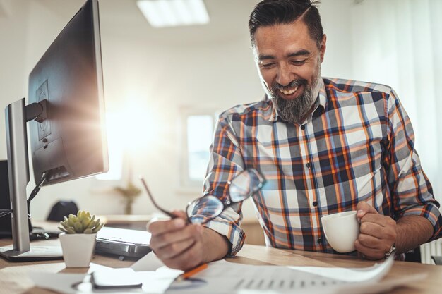 若いハンサムな成功したビジネスマンは、プロジェクトを分析し、コーヒーを飲みながら、オフィスで働いています。