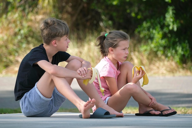 Young handsome teenager boy and pretty girl eating tasty ripe banana snacking outdoors on summer day