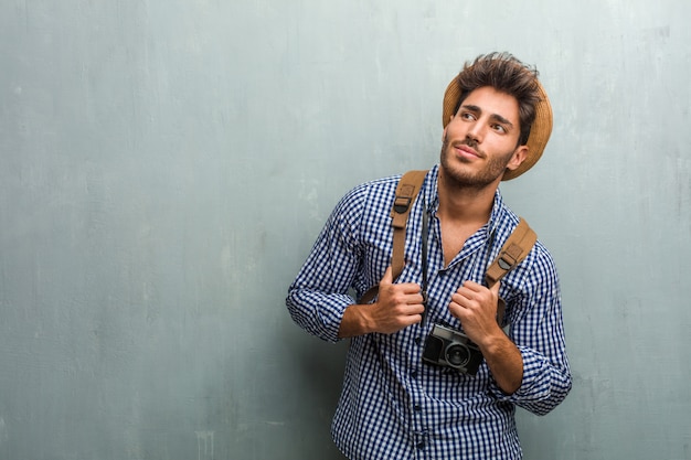 Young handsome traveler man wearing a straw hat, a backpack and a photo camera looking up