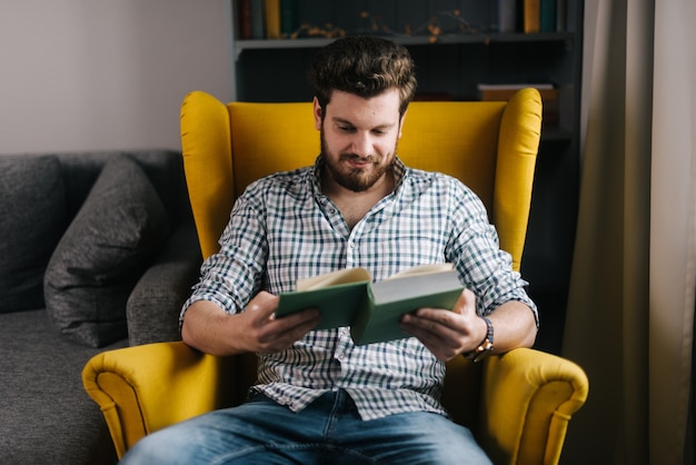 Фото Молодой красивый молодой человек с бородой сидит на желтом стуле и читает книгу