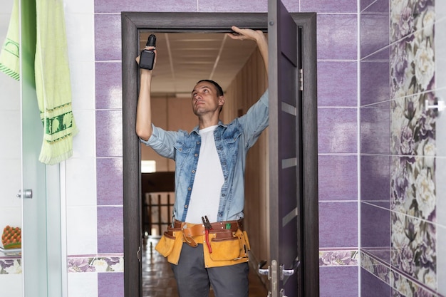 Young handyman installing a white door with an electric hand drill in a room.