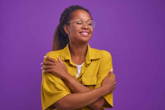 Young happy african american woman hugs herself stands in purple studio