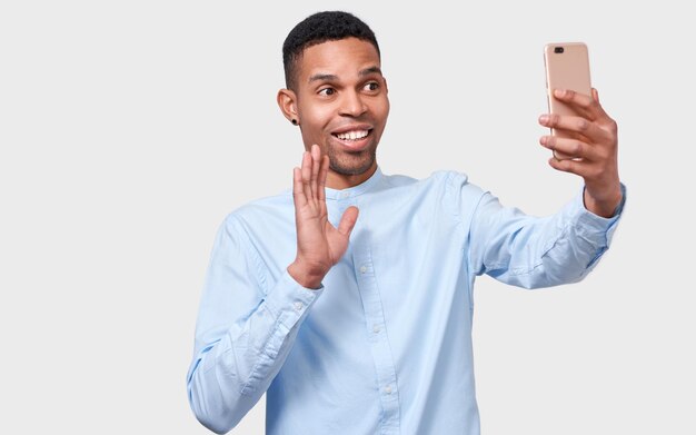 Young happy African American young man holds modern smart phone speaking online and blogging Handsome Afro male smiling broadly sending hello gesture to his friend via Internet People technology