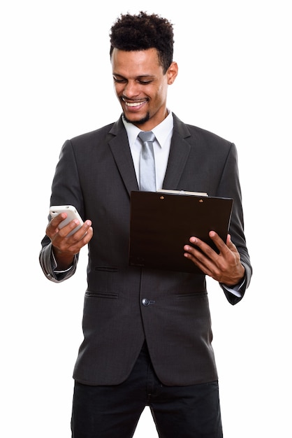 Young happy African businessman smiling while holding clipboard and using mobile phone