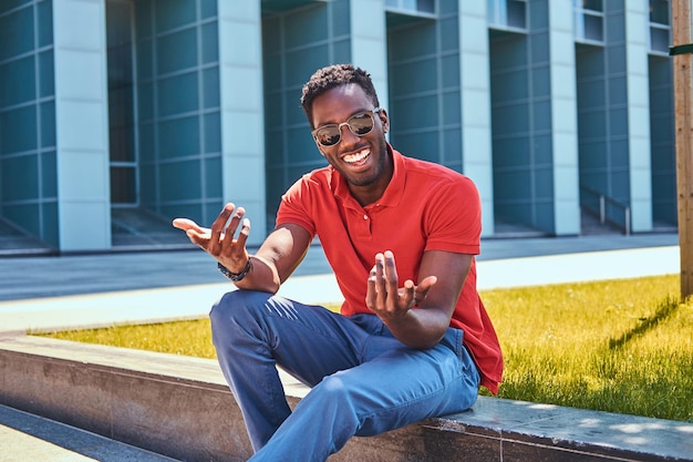 Il giovane uomo afroamericano felice in occhiali da sole è seduto sull'erba vicino al grattacielo.