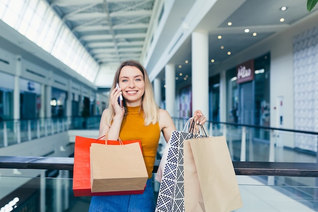 Foto giovane donna felice dopo lo shopping parlando al cellulare nel centro commerciale. shopaholic femminile gioioso soddisfatto con sacchetti di carta colorati regalo nelle mani. conversazione al cellulare. gioia sorridente stand ang sinceramente racconta