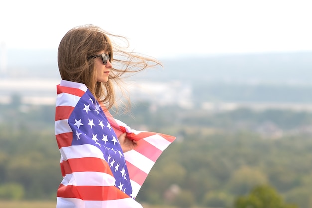 Молодая счастливая американская женщина с длинными волосами, держащая размахивая на ветру Национальный флаг США на ее плечах, расслабляющихся на открытом воздухе, наслаждаясь теплым летним днем.