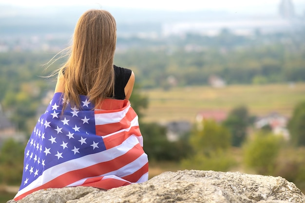 Young happy american woman with long hair holding waving on wind USA national flag on her sholders relaxing outdoors enjoying warm summer day.