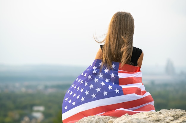 Young happy american woman with long hair holding waving on wind USA national flag on her sholders relaxing outdoors enjoying warm summer day