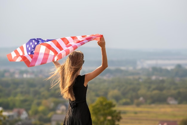 Young happy american woman with long hair raising up waving on wind usa national flag in her hands relaxing outdoors enjoying warm summer day.