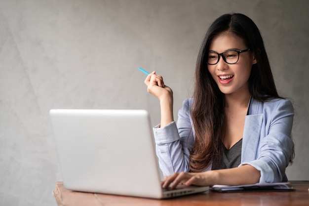 Young happy Asian businesswoman in blue shirt working from home and use a computer laptop and thinking idea for her business