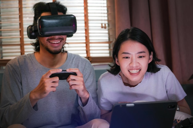 Young happy asian couple playing video games virtual reality glasses in living room. Cheerful people having fun with new trends technology. Man with hands up wearing the virtual reality goggles.