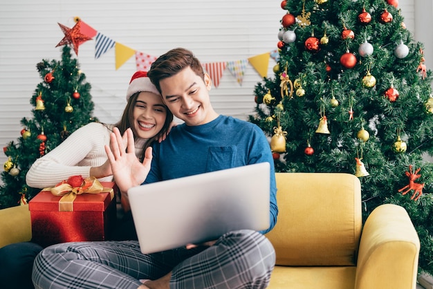 Young happy asian couple sitting together video call on a\
laptop with friends or parents thanks for a gift received during\
the christmas holiday with a decorated christmas tree in the\
background