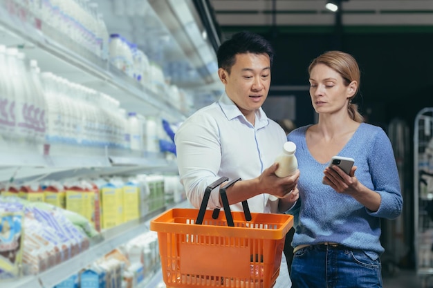 Young happy asian couple using smartphone in supermarket with shopping cart choosing products while