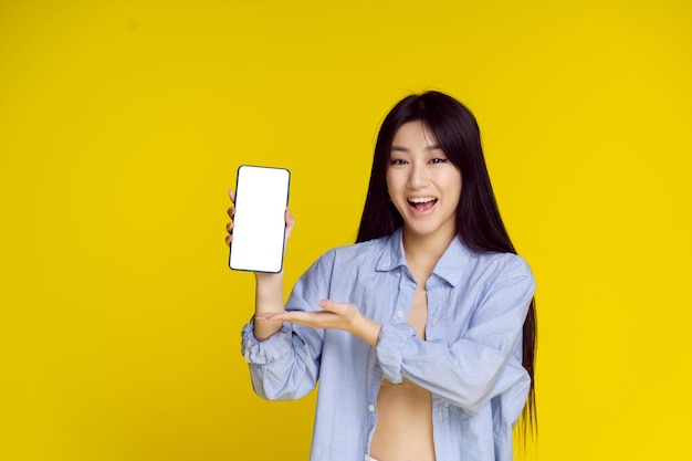 Young happy asian girl holding smartphone showing a white empty screen and exciting to win isolated on yellow great offer