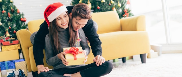 A young happy Asian man blowing a party whistle in the back at his girlfriend wearing a Santa Claus hat holding a received present at home with a Christmas tree in the background