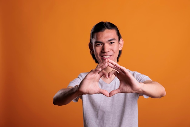 Young happy asian man feeling love, showing heart shape with\
fingers, focus on hands. smiling cheerful teenager making\
appreciation sign, romance symbol, looking at camera