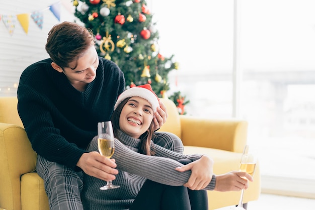 Il giovane uomo asiatico felice sorride e ride alle spalle della sua ragazza che indossa un cappello di babbo natale mentre beve champagne a casa con un albero di natale sullo sfondo