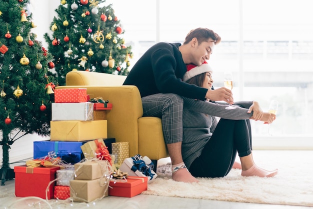 若い幸せなアジアの驚きとバック グラウンドでクリスマス ツリーと自宅でシャンパンを飲みながらサンタ クロースの帽子をかぶっている彼のガール フレンドの後ろで笑うコピー スペース イメージ