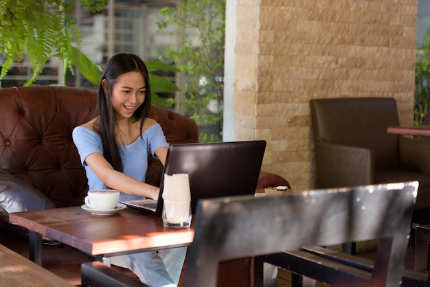 Young happy Asian teenage woman smiling at outdoor coffee shop while using laptop