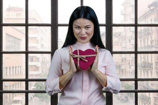 Молодая счастливая азиатка, держащая подарочную коробку в форме сердца обеими руками