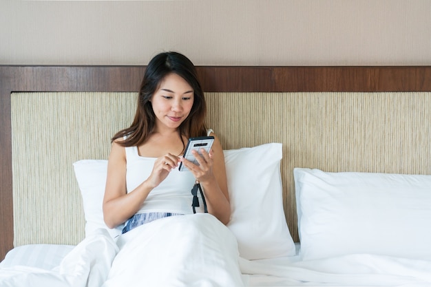 ベッドに座って携帯電話を使用して若い幸せなアジアの女性