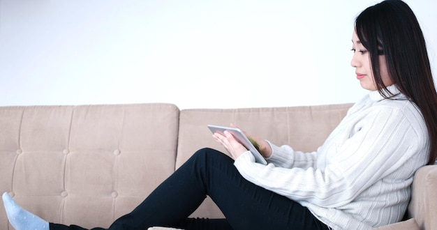 디지털 태블릿을 사용하여 소파에 앉아 있는 젊은 행복한 아시아 여성