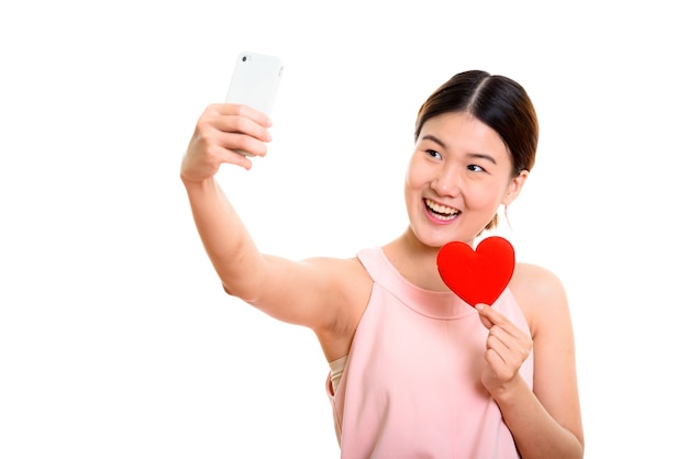 若い幸せなアジアの女性笑顔と赤いハートを保持しながら携帯電話で自分撮り写真を撮る