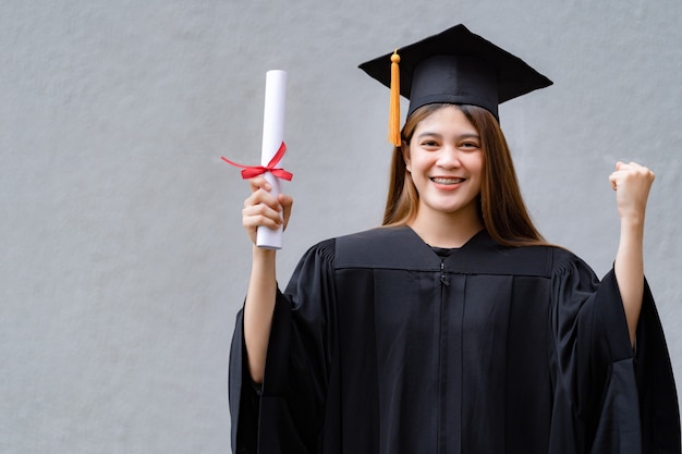 A young happy asian woman university graduate in graduation\
gown and mortarboard holds a degree certificate celebrates\
education achievement in the university campus. education stock\
photo