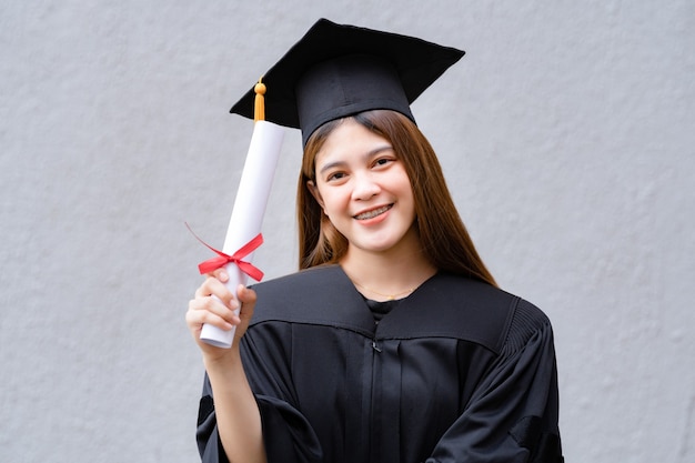 A young happy asian woman university graduate in graduation\
gown and mortarboard holds a degree certificate celebrates\
education achievement in the university campus. education stock\
photo