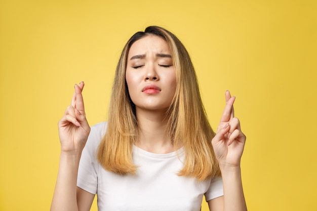 Young happy asian woman with fingers crossed on yellow background